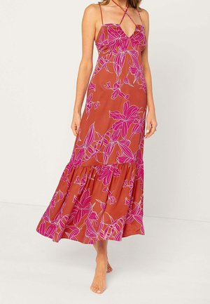 Femme portant une robe longue couleur rouille avec un imprimé floral rose et de fines bretelles croisées, debout pieds nus sur un fond uni.