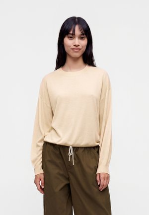 Jeune femme aux longs cheveux foncés portant un chemisier beige à manches longues et un pantalon vert olive, debout devant un fond clair uni.