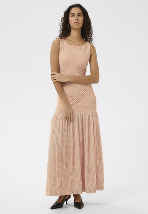 Frau mit lockigem Haar, die ein ärmelloses, strukturiertes Maxikleid in Pfirsichfarbe und schwarze spitze High Heels trägt, mit verschränkten Armen vor einem weißen Hintergrund steht.