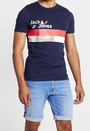 Mężczyzna w granatowej koszulce Jack & Jones z czerwonymi i białymi paskami, w połączeniu z jasnoniebieskimi podwiniętymi szortami jeansowymi.