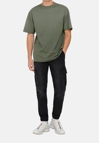 Olivgrünes T-Shirt mit kurzen Ärmeln, lockere Passform, kombiniert mit schwarzen Cargo-Hosen mit Seitentaschen und weißen Sneakern mit mehrfarbigen Akzenten.