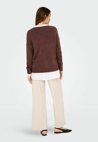 Donna vista di spalle che indossa un maglione marrone sopra una camicia bianca, con pantaloni larghi color crema e scarpe slingback nere su uno sfondo neutro.