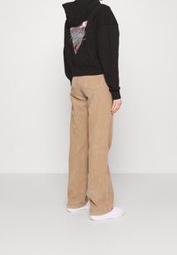Sudadera con capucha negra con gráficos en la parte posterior, combinada con pantalones de pana beige de pierna amplia y zapatillas blancas. Texturas suaves visibles.