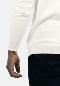 Witte gebreide trui met een geribde manchette, gedragen met een donkere broek. Focus op de textuur en pasvorm rond de arm- en taillezone.