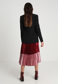 Sort skræddersyet blazer parret med en plisseret nederdel i nuancer af rød og pink. Nederdelen har en skinnende overflade og lagdelt design.