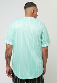 Gai minta zaļa džerzeja blūze ar vertikālām baltām svītrām, īsām piedurknēm, noapaļotu apakšmalu un mīkstu tekstūru, demonstrējot ikdienišķu, brīvu piegriezumu.