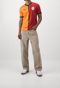 Camiseta de fútbol Puma con un diseño dividido en rojo y naranja, mangas cortas, cuello en V y un logo del club. Pantalones beige y zapatillas blancas complementan el conjunto.