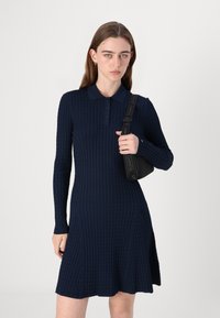 Marineblå ribbestrikket kjole med lange ermer, polo-krage, knappestengning, sidespalte, kombinert med en liten svart skulderveske.