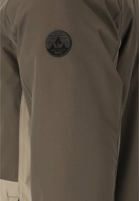 Khaki-jacka med en slät tygtextur, med en svart cirkulär logotyp-patch och kontrasterande ljusare tyg på sidan.