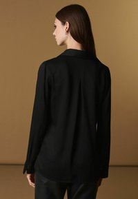 Sort langærmet skjorte med fold bagpå, krave og knapdetaljer ved cuffen. Glat tekstur, blødt stof og skræddersyet design.