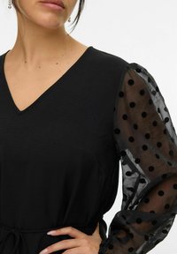 Zwarte jurk met een halslijn in V-vorm, voorzien van doorschijnende pofmouwen versierd met zwarte stippen. Glad materiaal met een gestructureerde uitstraling.