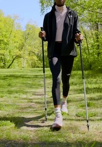 Planinarske štake drži osoba u crnoj jakni i ružičastom topu, obučena u crne tajice i svijetlo ružičaste cipele, koja hoda po travnatoj stazi.