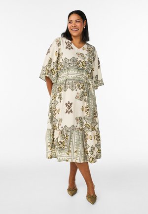 Žena sa usmieva, oblečená v krémových midi šatách s zelenými kvetinovými a geometrickými vzormi a olivovo zelenými špicatými balerínkami, stojaca na bielom pozadí.