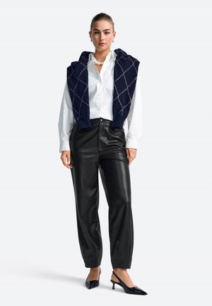 Pulover cu model argyle în nuanțe de navy, așezat peste o cămașă albă cu nasturi, asociat cu pantaloni din piele negri și pantofi cu vârf ascuțit negri.