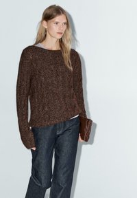 Maglione marrone a maglia con un motivo testurizzato, vestibilità rilassata e maniche lunghe, abbinato a pantaloni in denim scuro e una borsa a mano marrone.