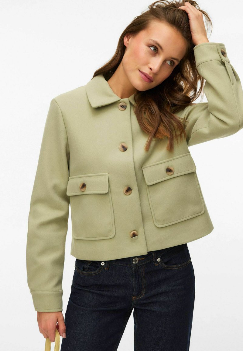 Femme portant une veste boutonnée vert clair avec de grandes poches, un jean foncé, tenant un sac beige et se touchant les cheveux.