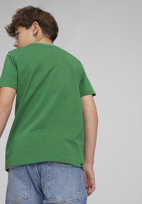 Camiseta de algodón verde con mangas cortas y cuello redondo. Presenta una sutil costura en contraste alrededor del cuello y una pequeña etiqueta de la marca en el dobladillo.