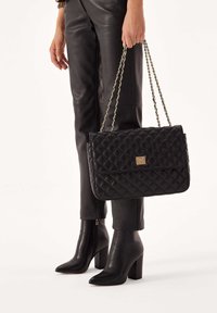 Borsa a tracolla in pelle nera trapuntata con chiusura in hardware dorato e tracolla a catena. Abbinata a pantaloni in pelle nera e stivali con tacco alto.
