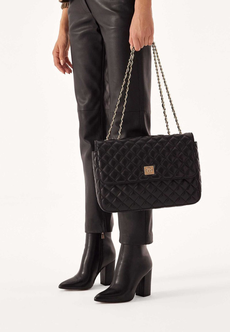 Borsa a tracolla in pelle nera trapuntata con chiusura in hardware dorato e tracolla a catena. Abbinata a pantaloni in pelle nera e stivali con tacco alto.
