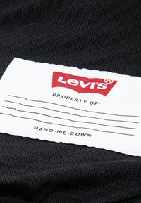 Crna tkanina s bijelom etiketom. Etiketa sadrži Levi's logo u crvenoj boji, granicu svojine i tekst koji glasi "HAND-ME-DOWN."