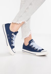 Baskets basses en toile bleu marine avec embout blanc en caoutchouc. Elles présentent des lacets rayés, des œillets métalliques et une semelle intermédiaire blanche avec des rayures bleues.