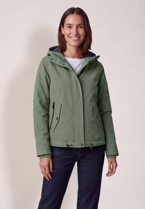 Femme souriante portant une veste à capuche verte par-dessus un haut blanc et un jean foncé, debout devant un fond clair uni.