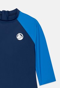 Marine og blå langermet rash guard med en tettsittende design, høy krage og en hvit seilbåtlogo på brystet. Jevn tekstur.