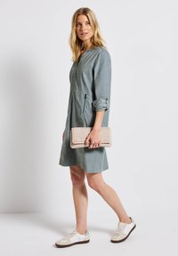 Kvinna som går i en grå knälång klänning med uppvikta ärmar, vita sneakers och håller en beige vävd clutchväska vid sin sida.