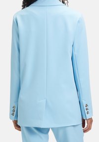 Blazer tailleur bleu clair, avec une fente unique à l'arrière et trois boutons dorés en accent sur la manche, confectionné dans un tissu lisse.