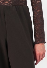 Brune, høy midje bukser laget av glatt stoff, kombinert med en gjennomsiktig blonde topp med lange ermer som har et blomstermønster langs ermene.