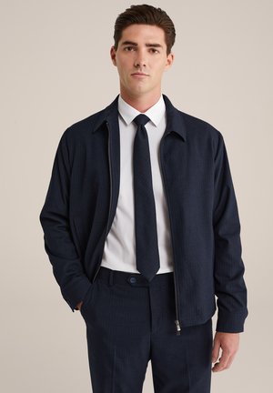 Veste bleu marine à rayures en biais avec fermeture éclair à l'avant, associée à une chemise blanche et une cravate foncée. Le tissu est texturé et lisse, mettant en avant une coupe moderne.