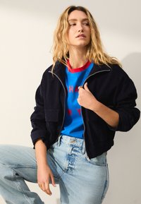 Chaqueta bomber de pana azul marino con cierre de cremallera y bolsillos delanteros, usada sobre una camiseta azul con detalles rojos y texto, combinada con vaqueros claros.