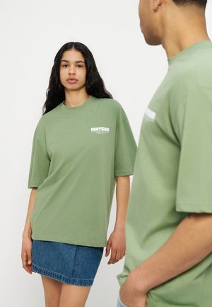 Zöld, túlméretezett pólót és farmer szoknyát viselő nő a kamerába néz, egy hozzá illő zöld pólót viselő férfi áll előtérben, és rá néz.