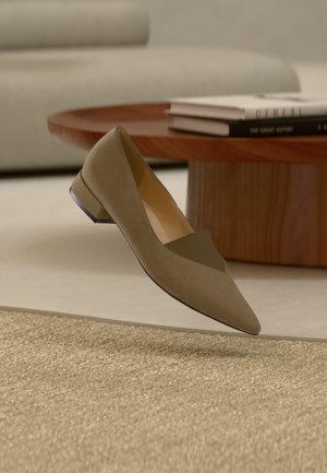 Singola scarpa piatta in camoscio verde oliva con tacco basso, posizionata sopra un tappeto beige intrecciato, tavolo di legno e pila di libri sullo sfondo.