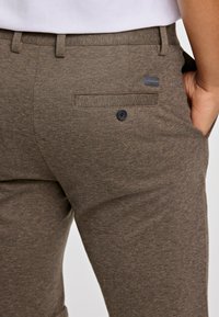 Bruine broek van textiel met een structuur, voorzien van een achterzak met een zwarte knop en subtiele branding op de tailleband.