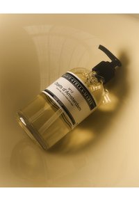 Bottiglia di sapone liquido in vetro trasparente con pompa nera, etichettata "Objets d'Amsterdam." Liquido giallo visibile. Design moderno e minimalista.
