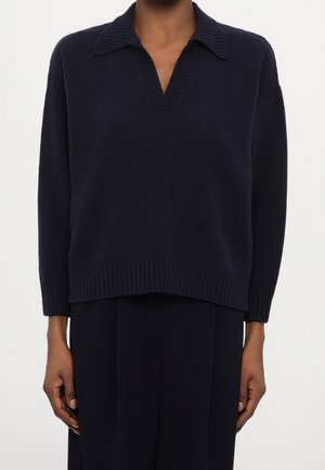 Persona con un suéter de punto azul marino oscuro con un cuello ancho y escote en V, combinado con pantalones negros sueltos, de pie frente a un fondo liso.