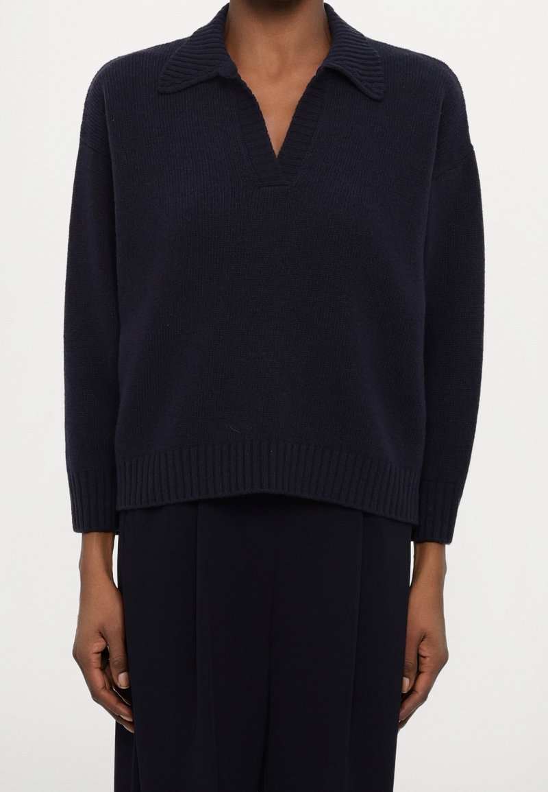 Personne portant un pull en tricot bleu marine foncé avec un large col et un col en V, associé à un pantalon noir ample, debout devant un fond uni.