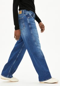 Wijd uitlopende blauwe spijkerbroek in een vervaagde was, met een hoge taille en zichtbare stiksels, gecombineerd met crèmekleurige schoenen.