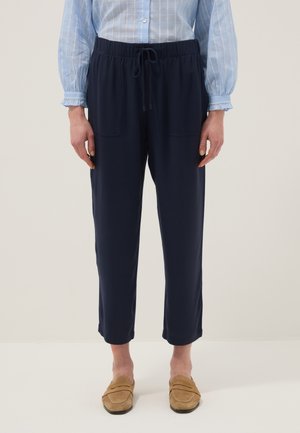 Femme portant un pantalon à cordon de serrage bleu marine, une chemise à manches longues à carreaux bleus et des mocassins beiges, debout devant un fond uni.