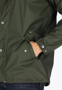 Green waterproof jacket made of smooth material, featuring silver snap buttons and side pockets, with a relaxed fit.