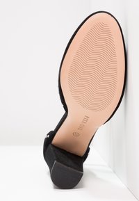 Svart mocka högklackad sko med en slät, texturerad sula, som har rundad tå och dekorativa detaljer på sidan.