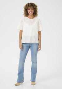 Blusa bianca ricamata con maniche a sbuffo corte, caratterizzata da motivi ad occhietto e un collo rotondo, abbinata a jeans a zampa d'elefante blu chiaro.