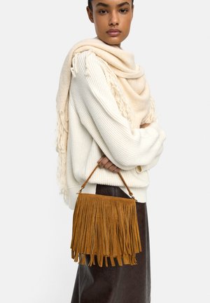 Kobieta ubrana w kremowy sweter z dzianiny, pasujący szalik z frędzlami, ciemną spódnicę, trzymająca brązową zamszową torebkę z frędzlami za uchwyt na jednolitym tle.