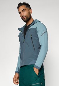 Kék-szürke kapucnis kültéri dzseki világosabb kontrasztos panelek kialakítással. Cipzáras mellzsebbel és textúrált anyaggal rendelkezik. Lélegző dizájn.