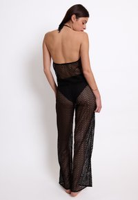 Tuta nera all'uncinetto con motivo a zigzag, top senza spalline e design a gamba larga. Presenta una texture trasparente e un dettaglio sulla schiena aperta.