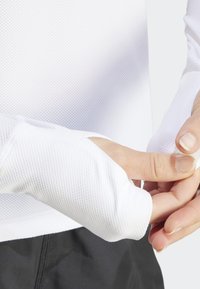 Biała, długosleeve'owa bluzka sportowa z siateczkową fakturą i otworami na kciuki; noszona z czarnymi spodami, ręce dostosowują rękaw.