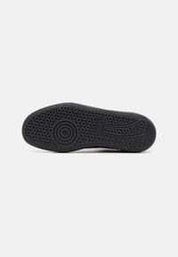 Semelle de chaussure noire avec motif de bande de roulement circulaire et rainurée et étiquette logo rectangulaire près du centre sur fond blanc.