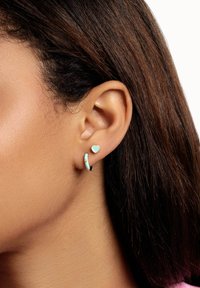 Orecchino a forma di cuore con bordi dorati e orecchino ad anello in turchese su un modello. L'anello presenta una finitura lucida, che si abbina al tono della pelle.
