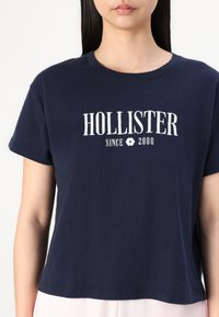 Námornícky modré bavlnené tričko s krátkymi rukávmi, s bielym textom "HOLLISTER" a ikonkou kvetu, okrúhly výstrih a voľný strih.